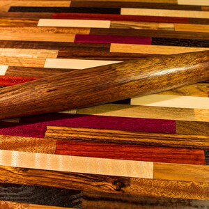 May include: A wooden rolling pin with a dark brown finish, resting on a colorful wooden cutting board made of various wood types.