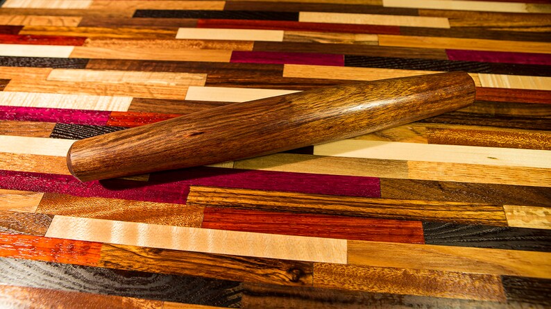 May include: A wooden rolling pin with a dark brown finish, resting on a colorful wooden cutting board made of various wood types.