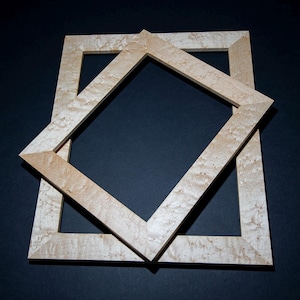 Puede incluir: Dos marcos de fotos de madera de color claro, uno posicionado diagonalmente sobre el otro, creando un efecto en capas. Los marcos tienen un patrón de vetas de madera natural sobre un fondo oscuro.