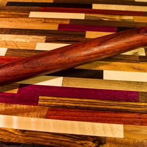 May include: A wooden rolling pin with a reddish-brown finish. The rolling pin is lying on a colorful wooden surface made of various wood types.