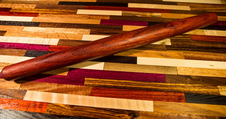 May include: A wooden rolling pin with a reddish-brown finish. The rolling pin is lying on a colorful wooden surface made of various wood types.