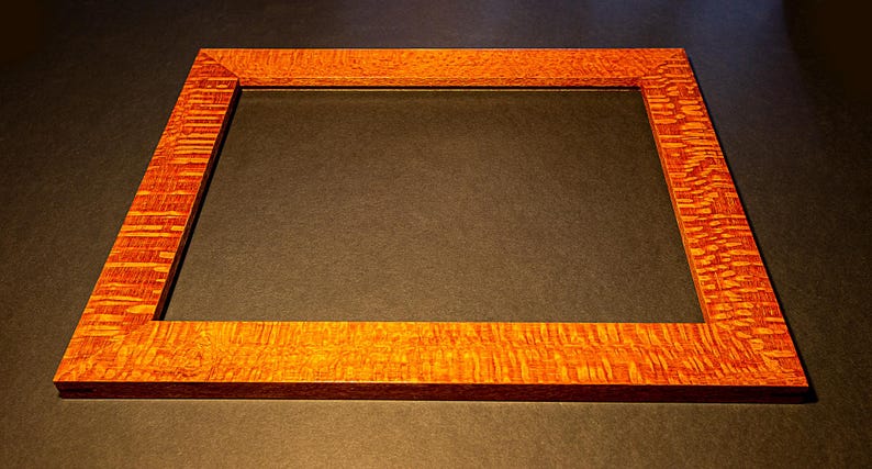 以下が含まれることがあります： 赤みがかった茶色の長方形の木製フォトフレーム。独特の木目模様が特徴です。フレームは空で、詳細な職人技と木の自然な美しさを際立たせています。写真やアートワークを収めるように設計されています。