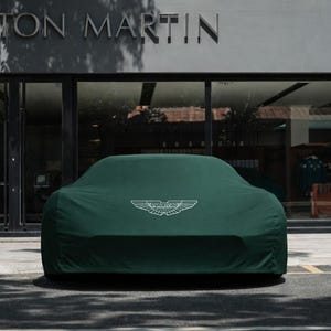 Op de afbeelding: Een donkergroene autohoes met het witte Aston Martin-logo, die een voertuig bedekt. De hoes staat voor een gebouw met de woorden "ASTON MARTIN" boven de ingang.