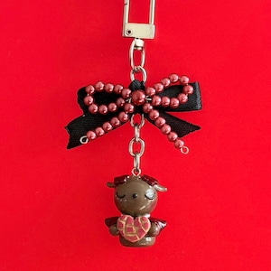 May include: A brown and red charm with a gold-coloured clasp. The charm features a small figurine with a heart-shaped design, suspended below a black bow adorned with red beads. The background is a solid red.
