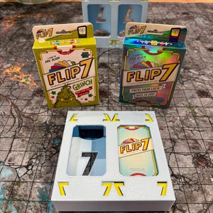 May include: A selection of "Flip 7" card game boxes in different colours, including green and iridescent, are shown. The game's title is displayed, along with the text "Press Your Luck! Race to 200!". A light blue box with the number 7 is also visible.