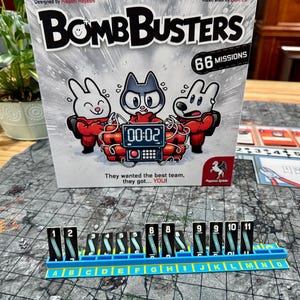 以下が含まれることがあります： ボードゲーム「Bomb Busters」は、漫画のキャラクターと「66 Missions」というテキストが描かれた箱が特徴です。 番号付きのタイルと文字が描かれた青いゲームボードが前景にあります。「They wanted the best team, they got... YOU!」というテキストも表示されています。
