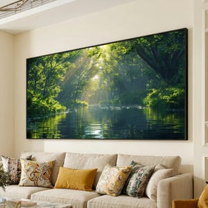 Può includere: Una grande stampa su tela incorniciata raffigurante una serena scena di foresta. La luce del sole filtra attraverso i lussureggianti alberi verdi, riflettendosi sull'acqua calma di un lago o fiume. L'opera d'arte è montata sopra un divano beige con cuscini decorativi.