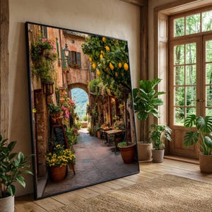 May include: A framed print depicting a European street scene. The image shows a narrow cobbled alleyway flanked by buildings, flowers, and trees with yellow fruit. A wooden door and plants are in the background.