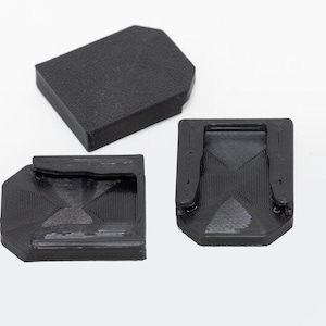 May include: Three black, geometric-shaped objects are displayed against a white background. The objects appear to be 3D-printed, with textured surfaces and angular designs. One is rectangular, and two have a more complex, angled form.