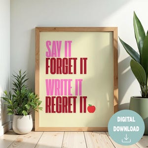 May include: A framed print with the text "SAY IT FORGET IT WRITE IT REGRET IT" in pink and red, with a red apple graphic. The print is in a light wood frame, with a digital download badge in the bottom right corner. The background is a light beige.