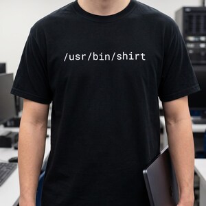 May include: A black short-sleeved t-shirt featuring the white text /usr/bin/shirt printed across the chest. The garment has a crew neckline and a standard fit, displaying a clear computer-themed command line path in a simple sans-serif font.