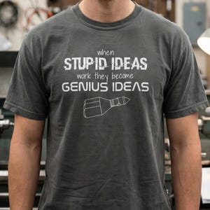 May include: Dark gray t-shirt with the text "when STUPID IDEAS work they become GENIUS IDEAS" in white. A white line drawing of a rocket is below the text. The shirt is a crew neck style.