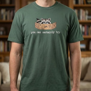 May include: Dark green t-shirt featuring a raccoon peeking out of a cardboard box. The box has the letters "DM" and the phrase "you can certainly try" printed on it. The shirt is made of a soft material.