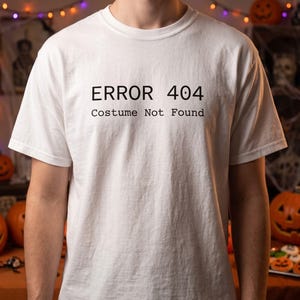 May include: A white short-sleeved t-shirt featuring black text printed on the chest. The text reads "ERROR 404" on the top line and "Costume Not Found" on the second line. The shirt has a crew neckline and a standard fit with a plain white fabric.
