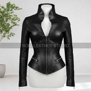 May include: A black leather jacket with a high collar and a zipper closure. The jacket has long sleeves, two zippered pockets, and the text "NOIR LEATHER STUDIO". The jacket is displayed on a mannequin.