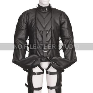 Puede incluir: Chaqueta de restricción de cuero negro con paneles acolchados, cuello alto y múltiples correas con hebillas. La chaqueta tiene mangas largas y se combina con sujeciones para las piernas a juego. El logotipo de Noir Leather Studio es visible.
