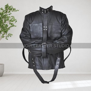 Può includere: Giacca in pelle nera con collo alto, due tasche frontali e cintura. La giacca ha cinghie per fissare le braccia e il corpo. Il testo "NOIR LEATHER STUDIO" è visibile sulla giacca.
