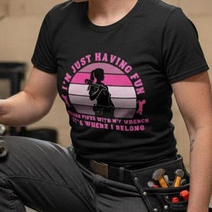 Puede incluir: Una mujer con una camiseta negra con el texto "I'M JUST HAVING FUN WITH MY WRENCH IT'S WHERE I BELONG" se arrodilla sosteniendo una llave inglesa. Está trabajando en tuberías de fontanería en un sótano.