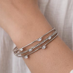 Könnte beinhalten: Drei silberfarbene Armreifen mit klaren, funkelnden Edelsteinen. Einer hat eine Reihe kleiner Steine, die anderen größere, ovale Edelsteine. Der Schmuck wird am Handgelenk vor weißem Hintergrund getragen.