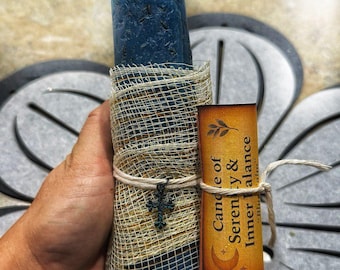 May include: A blue candle wrapped in a beige mesh and tied with twine, featuring a small cross charm. A label reads "Candle of Serenity & Inner Balance". The candle has a textured surface and a knotted wick.