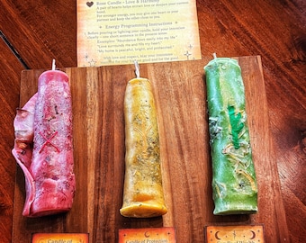 May include: Three handmade pillar candles in red, gold, and green, with accompanying cards. The red candle has a heart design, the gold candle has a cross design, and the green candle has a leaf design. The cards describe the candles' intentions: Love & Harmony, Protection & Cleansing, and Wealth & Prosperity.