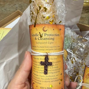 Könnte beinhalten: Eine Kerze mit der Aufschrift "Candle of Protection & Cleansing" und dem Text "Safeguard Light". Die Kerze ist in Luftpolsterfolie verpackt und mit Bindfaden verschnürt, mit einem dekorativen Kreuzanhänger. Goldkonfetti verziert die Verpackung.
