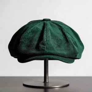 Puede incluir: Gorra de estilo newsboy de pana verde oscuro. La gorra se exhibe sobre un soporte metálico sobre un fondo blanco. El sombrero tiene una corona redondeada y una visera corta y rígida. La tela de pana tiene estrías verticales.