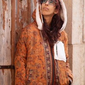 May include: An orange quilted jacket with a floral pattern and a white-lined hood. The jacket has a zip closure and a white pocket. The jacket is worn with light-coloured jeans. The image includes the text "vera luis".