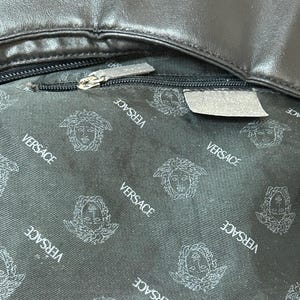 May include: Close-up of a black leather bag interior. The lining features a repeating pattern of the Versace logo in white. A silver zipper is partially open, revealing the interior. The bag's leather exterior is visible at the top.