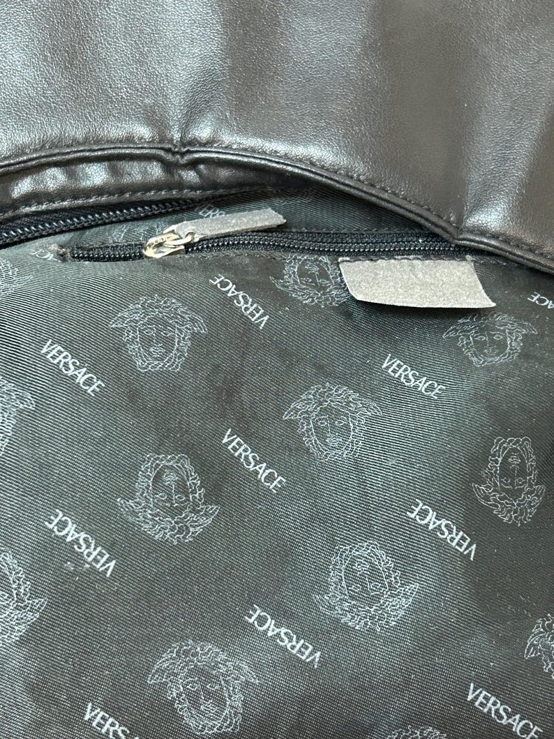 May include: Close-up of a black leather bag interior. The lining features a repeating pattern of the Versace logo in white. A silver zipper is partially open, revealing the interior. The bag's leather exterior is visible at the top.