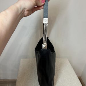 May include: Black leather handbag with a single handle. The handle is black with silver-tone metal accents and a chain detail. The bag is held up by a hand.
