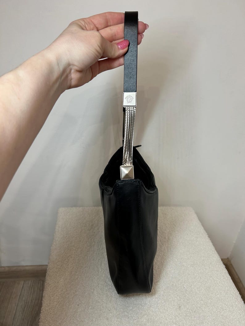 May include: Black leather handbag with a single handle. The handle is black with silver-tone metal accents and a chain detail. The bag is held up by a hand.