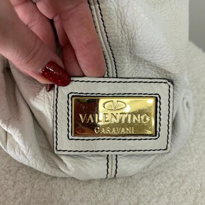 May include: White leather handbag with black stitching and a gold Valentino Garavani logo plate. The bag has a soft, textured appearance. The stitching is visible along the seams and around the logo plate.