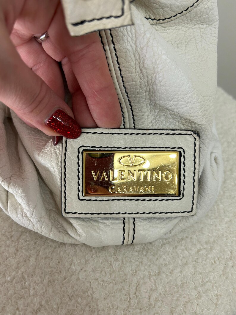 May include: White leather handbag with black stitching and a gold Valentino Garavani logo plate. The bag has a soft, textured appearance. The stitching is visible along the seams and around the logo plate.