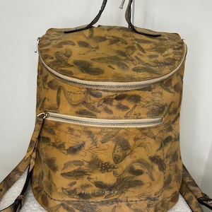 Può includere: Uno zaino in pelle color cuoio con una stampa marrone scuro di piume e esche da pesca. La borsa presenta una parte superiore arrotondata, un manico, due spallacci regolabili e due scomparti con cerniera. Il testo PLINIO VISONA è stampato sul davanti.