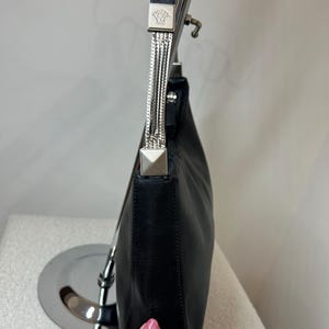 May include: Black leather shoulder bag with silver-tone hardware. The bag features a chain strap and a square metal accent with a Medusa head logo. The bag is displayed on a silver stand.