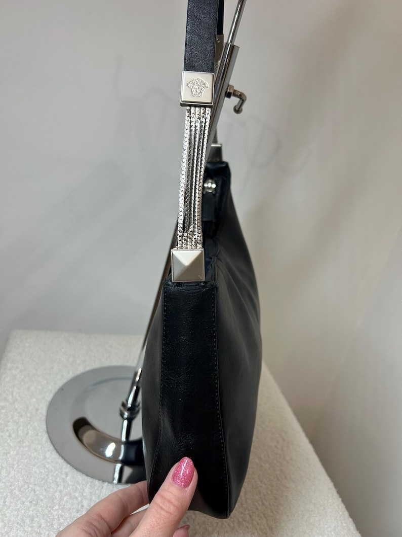 May include: Black leather shoulder bag with silver-tone hardware. The bag features a chain strap and a square metal accent with a Medusa head logo. The bag is displayed on a silver stand.