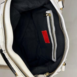 May include: White leather handbag with black accents and gold-tone hardware. The interior is black with a red rectangular tag. The bag features a zipper closure and a textured exterior.