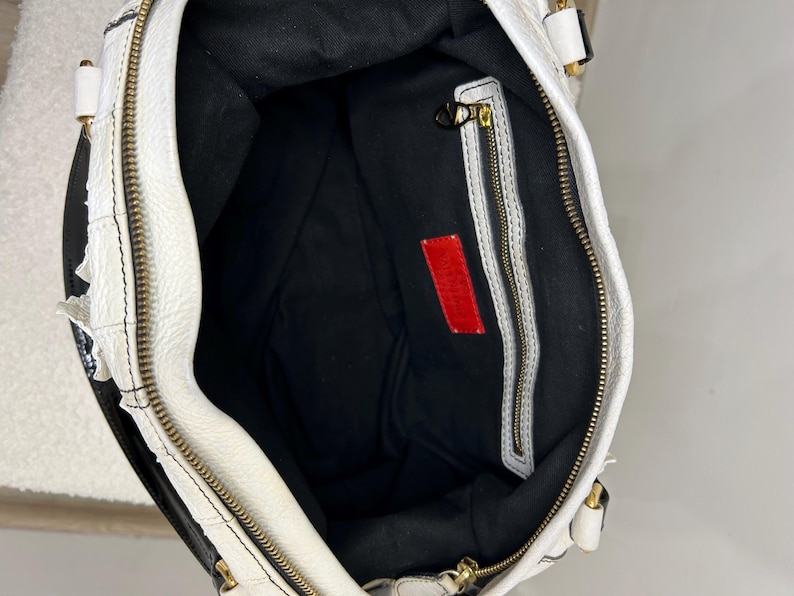 May include: White leather handbag with black accents and gold-tone hardware. The interior is black with a red rectangular tag. The bag features a zipper closure and a textured exterior.