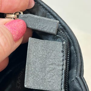 May include: Close-up of a black leather bag with a silver zipper. The bag has a gray tag attached to the zipper. The tag is rectangular and has a textured surface. The word "VERSACE" is visible on the bag's interior.