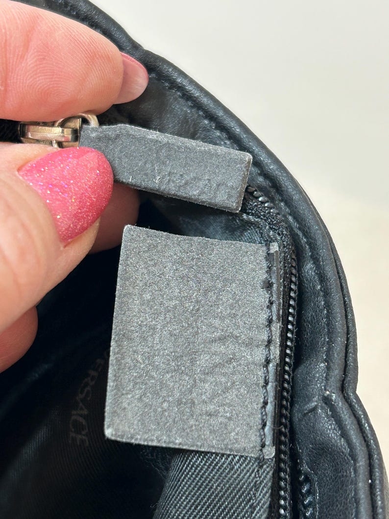 May include: Close-up of a black leather bag with a silver zipper. The bag has a gray tag attached to the zipper. The tag is rectangular and has a textured surface. The word "VERSACE" is visible on the bag's interior.