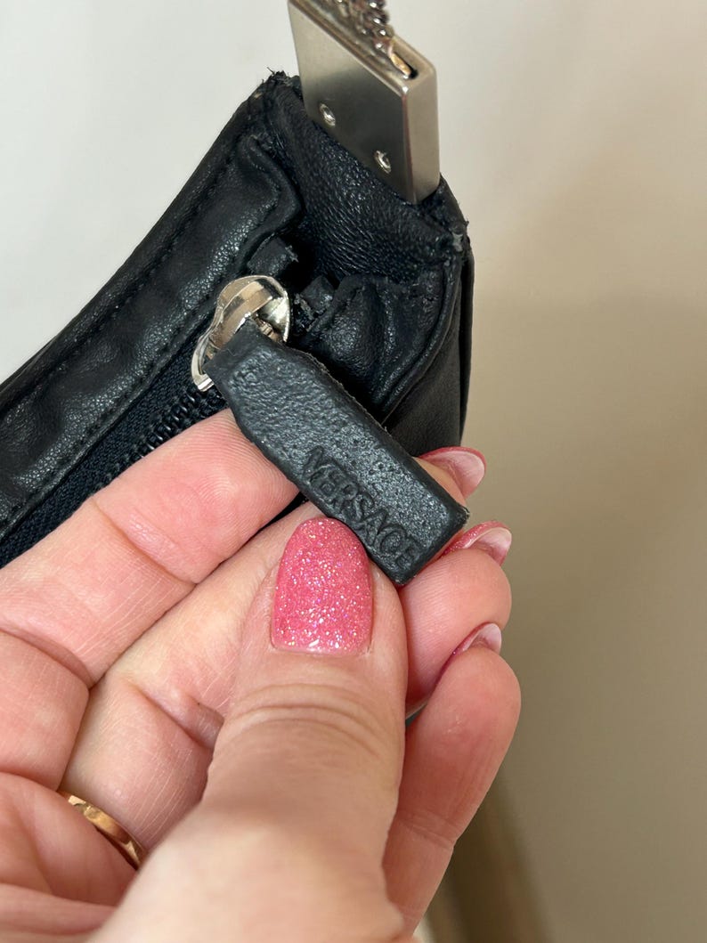 May include: Close-up of a black leather handbag with a silver zipper and chain strap. The zipper pull features the embossed brand name "VERSACE". The bag is held by a person.