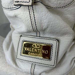 May include: White leather handbag with black stitching and a gold Valentino Garavani logo plate. The bag has a textured surface and a top handle with a gold-tone button.