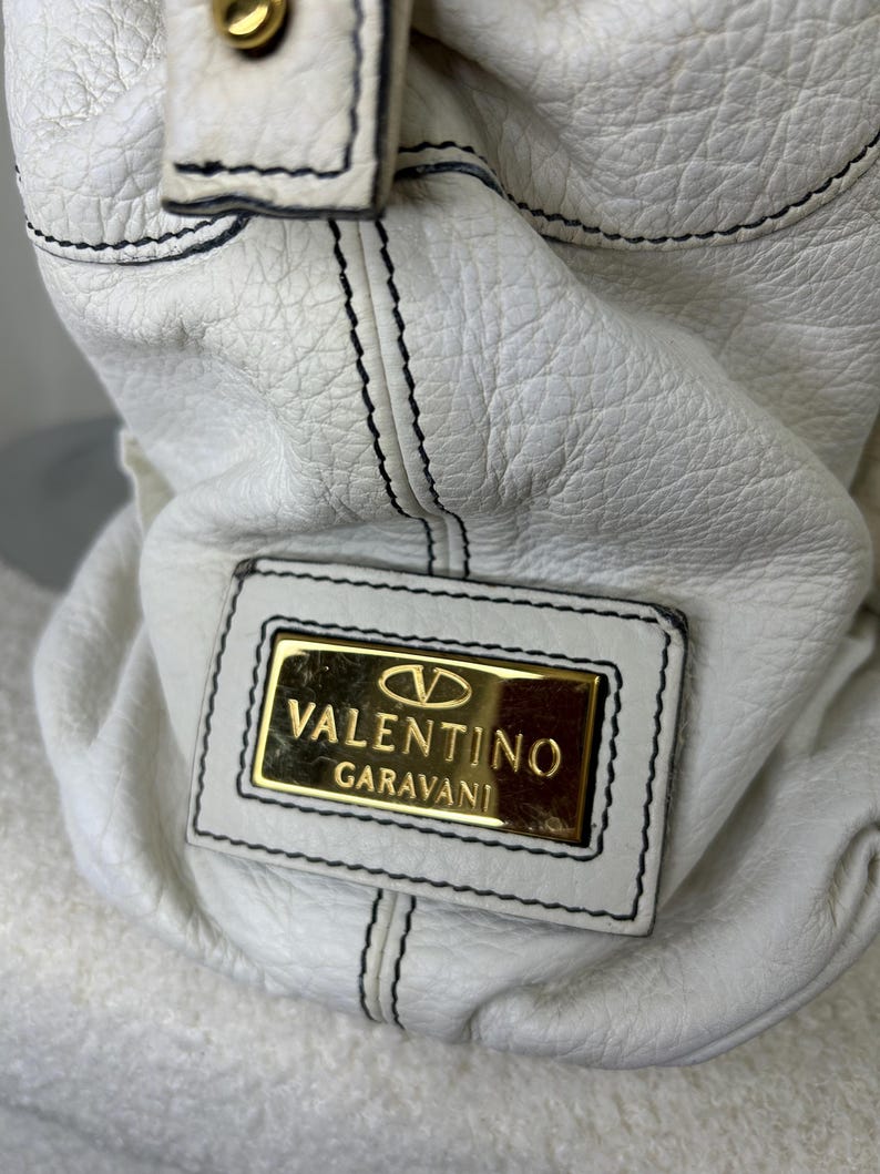May include: White leather handbag with black stitching and a gold Valentino Garavani logo plate. The bag has a textured surface and a top handle with a gold-tone button.