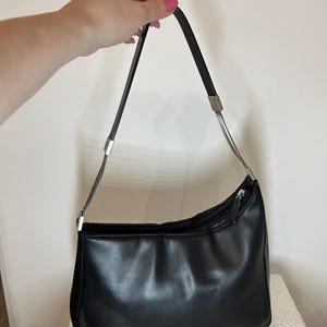 May include: Black shoulder bag with a curved design. The bag has a zipper closure and a silver chain detail on the strap. The bag is made of a smooth, shiny material.
