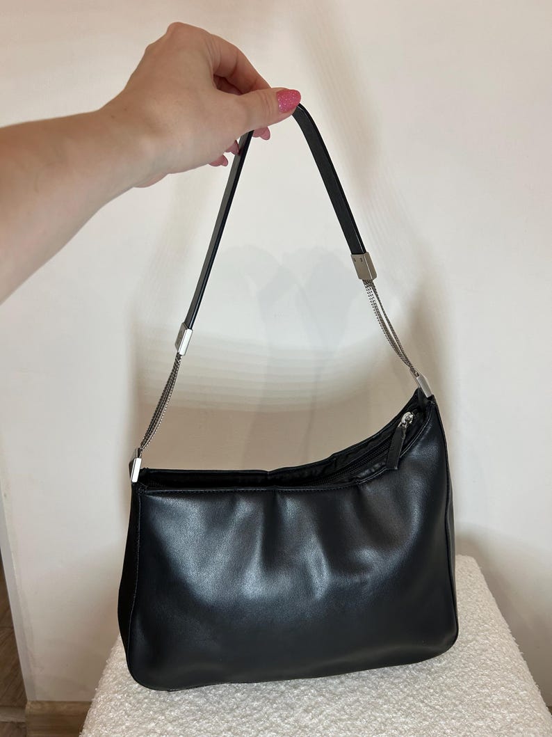 May include: Black shoulder bag with a curved design. The bag has a zipper closure and a silver chain detail on the strap. The bag is made of a smooth, shiny material.