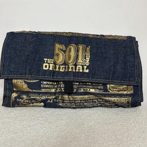 Op de afbeelding: Een donkerblauwe denim clutch met een overslag. De voorkant toont de tekst "501 THE ORIGINAL" in gouden letters. De clutch heeft een vintage uitstraling met gouden accenten en tekstdetails.