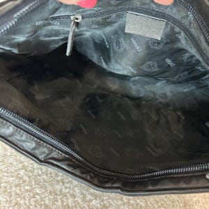 May include: Black leather handbag with a zipper closure. The interior lining is black with a repeating Versace logo pattern. A small, rectangular tag is visible inside the bag.