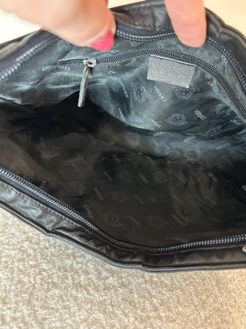 May include: Black leather handbag with a zipper closure. The interior lining is black with a repeating Versace logo pattern. A small, rectangular tag is visible inside the bag.
