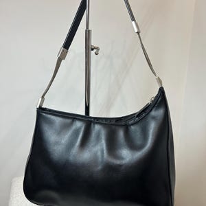 May include: Black shoulder bag with a curved design. The bag features a silver chain and black leather straps. The bag is displayed on a silver stand. The bag has a zipper closure.
